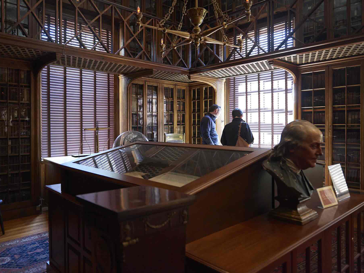 Visitors explore Pennsylvania Hospital's historic medical library, surrounded by floor to ceiling wooden bookcases, shuttered windows and exhibits, including a bust of Benjamin Franklin.
