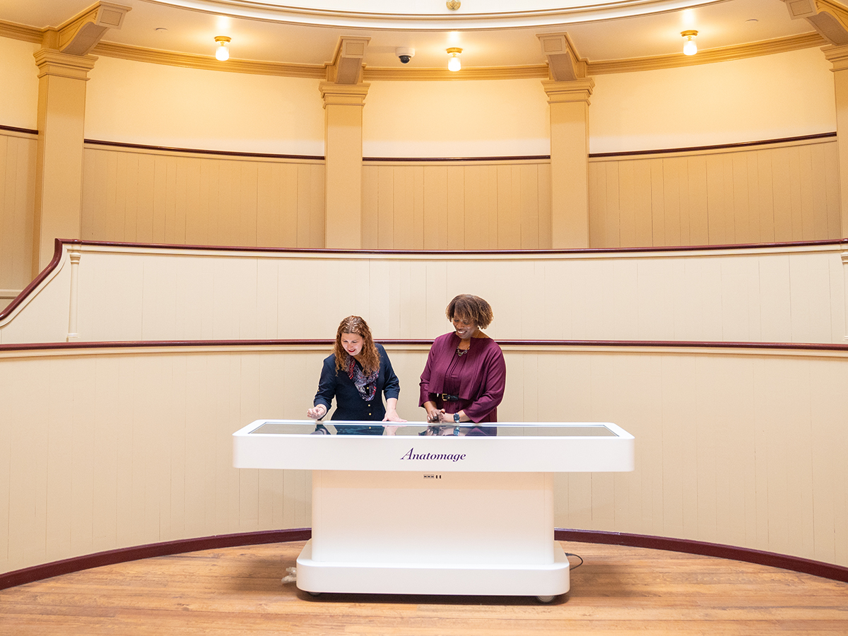 Two people use an interactive Anatomage table inside Pennsylvania Hospital's historic surgical amphitheater, surrounded by curved tiers once used for observing medical procedures.