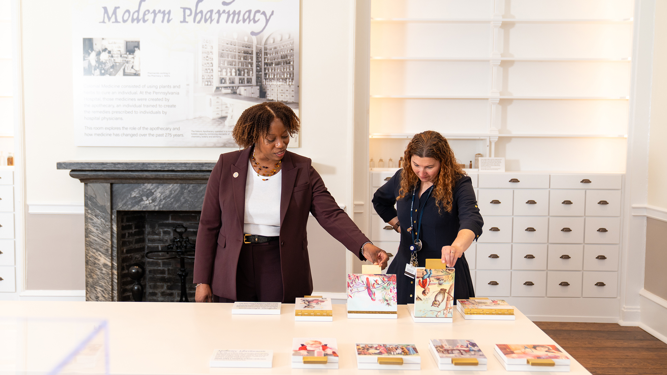 Two people examine an interactive display table in a museum exhibit exploring the evolution of pharmacy, with historic white cabinetry and black fireplace in the background.