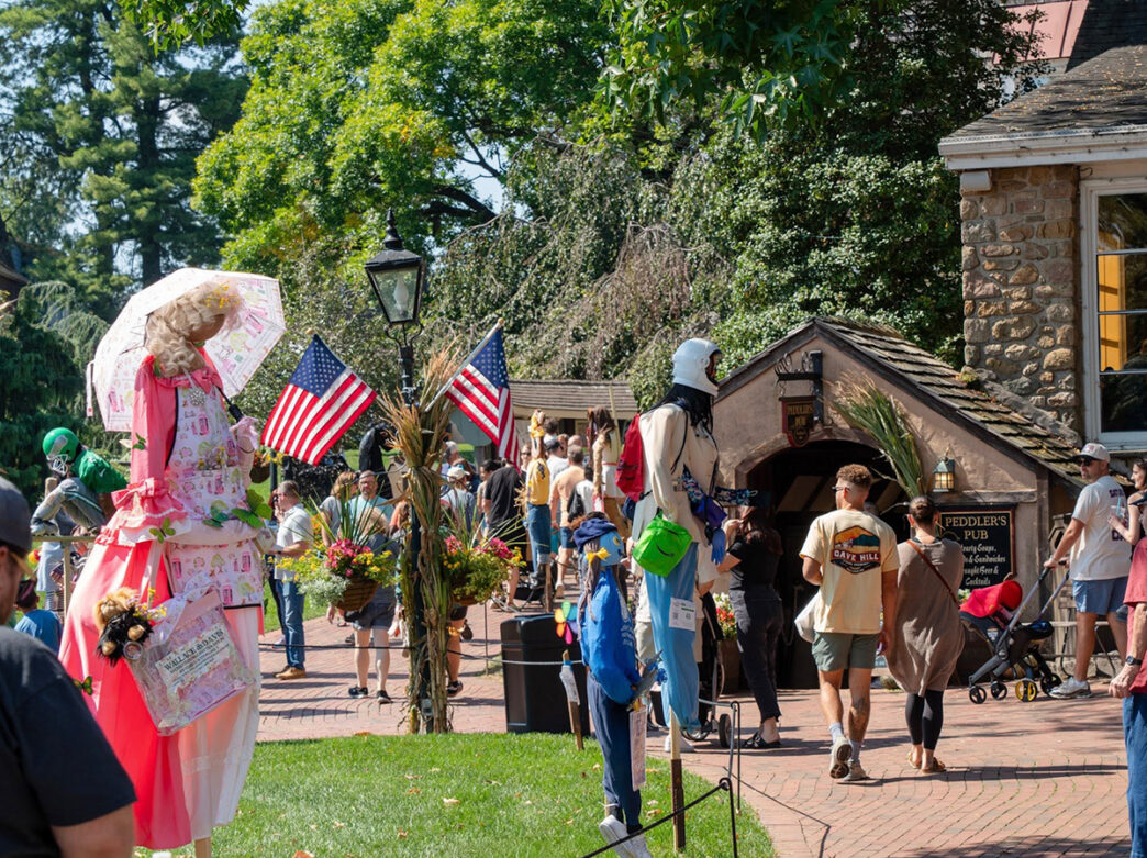 Visitors stroll through Peddler's Village during Scarecrows in the Village, with colorful scarecrows, fall decorations and American flags lines the brick walkway.