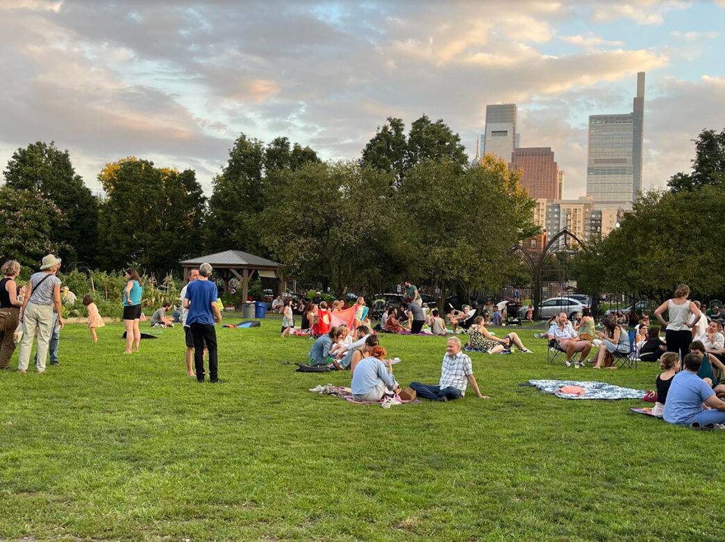 Durante el evento «Parks on Tap», grupos de personas se relajan sobre mantas y sillas de jardín repartidas por un césped, con el horizonte del centro de la ciudad asomándose más allá de los árboles.