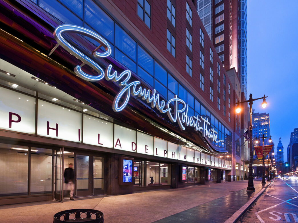 The exterior of Philadelphia's Suzanne Roberts Theatre at dusk, with its illuminated cursive sign and Philadelphia Theatre Company marquee stretching along the building exterior.