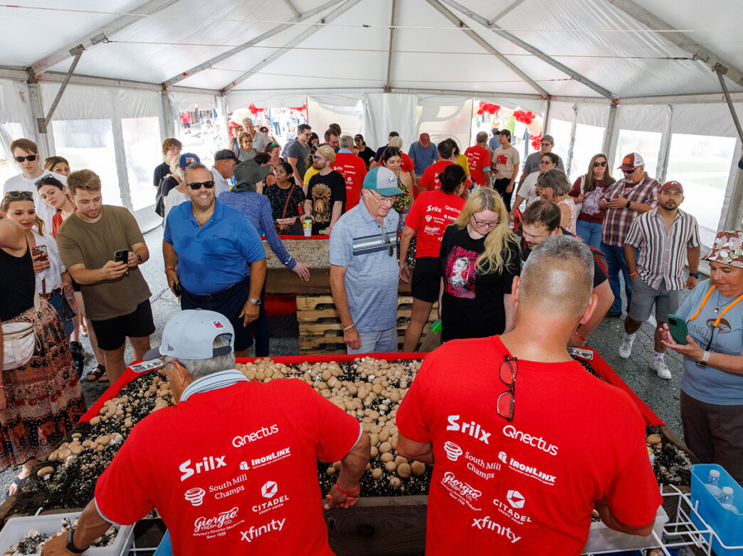 Los asistentes al festival se agolpan alrededor de mesas repletas de hongos dentro de una gran carpa blanca, mientras los vendedores, vestidos con camisetas rojas, atienden a los asistentes.
