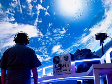 Lors d'un spectacle immersif du Philadelphia Fringe Festival, un artiste portant un casque audio se tient devant une grande projection de la Terre, au cœur d'un espace sombre baigné d'une lumière bleue.