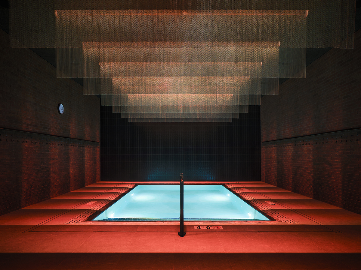 Une piscine d'eau froide d'intérieur aux reflets chatoyants, située dans une pièce baignée d'une lumière rouge chaleureuse et agrémentée d'une installation suspendue au plafond.