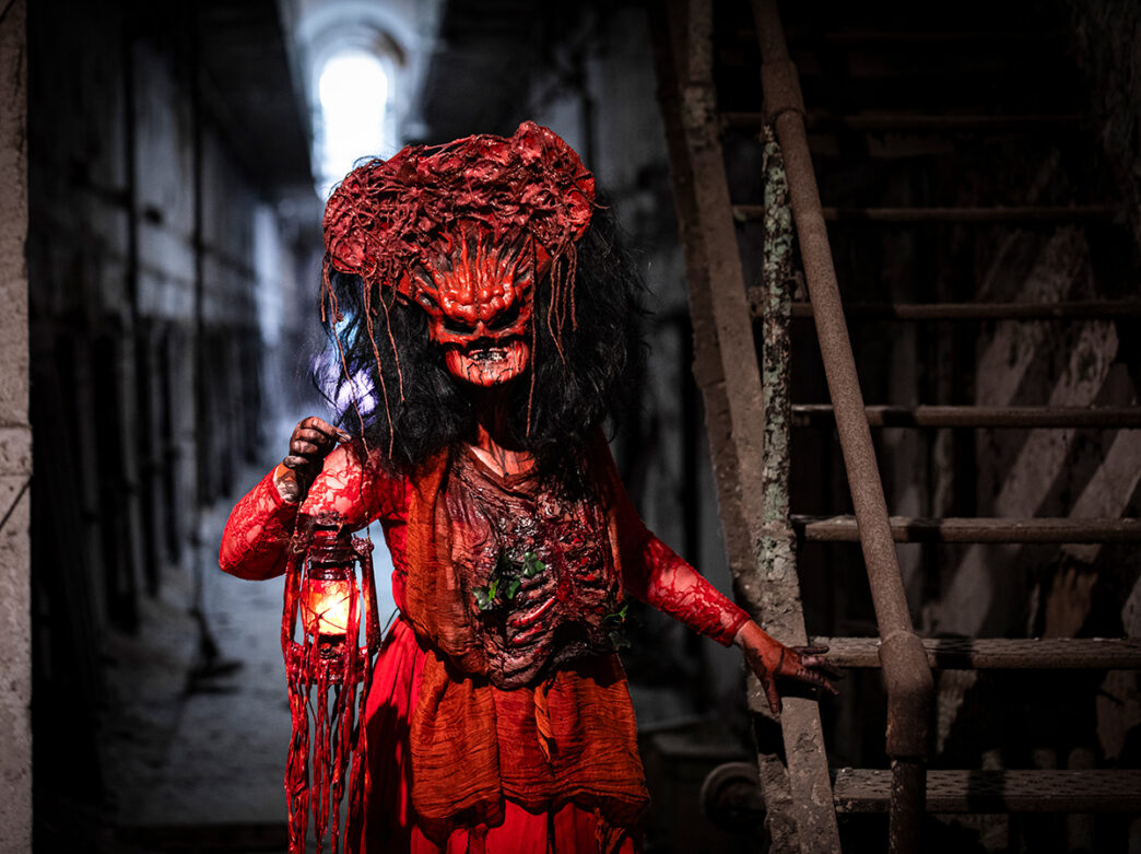 A costumed performer with a gory red mask, tattered red clothing and a glowing lantern lurks in a dark corridor at Eastern State Penitentiary.