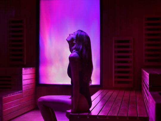 A woman relaxes in an infrared sauna illuminated with vibrant pink and purple light, surrounded by wooden benches and sauna panels.
