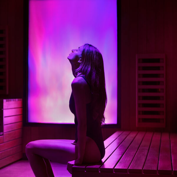 Une femme se détend dans un sauna infrarouge baigné d'une lumière rose et violette éclatante, entourée de bancs en bois et de panneaux de sauna.
