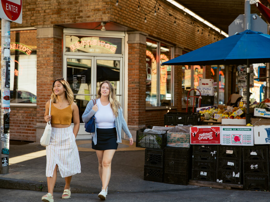 Two women stroll past Esposito's in the Italian Market, with stacked produce crates and an outdoor fruit and vegetable stand lining the sidewalk.