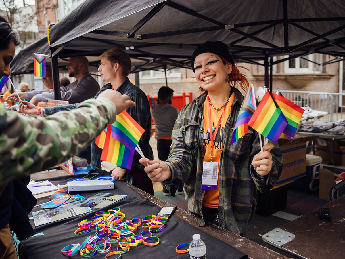 A smiling Ourfest vendor holds out Pride flags at a festival booth while a festivalgoer grabs a mini flag.