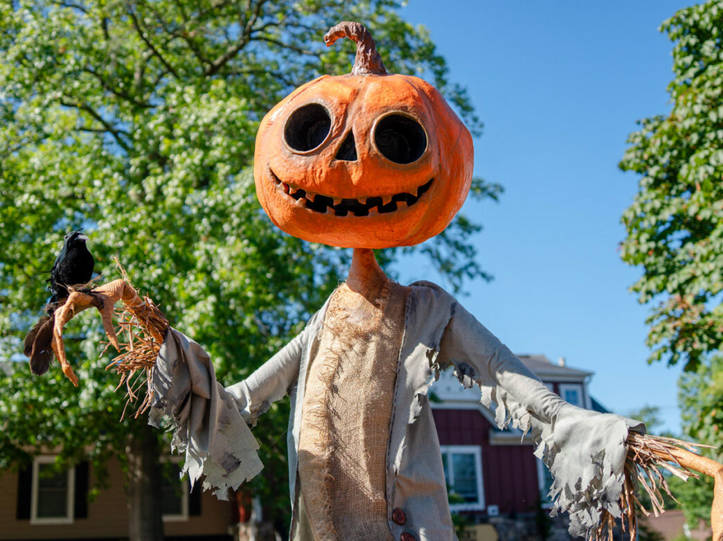 Un espantapájaros con forma de calabaza de Halloween de color naranja, con ropa hecha jirones y un cuervo negro posado en su mano, sonríe en Peddler’s Village.