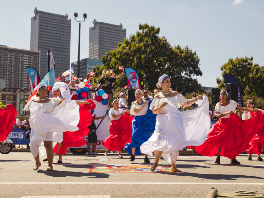 Dancers in flowing red, white and blue dresses perform in the Puerto Rican Day Parade on a car free street as spectators and parade participants gather behind them