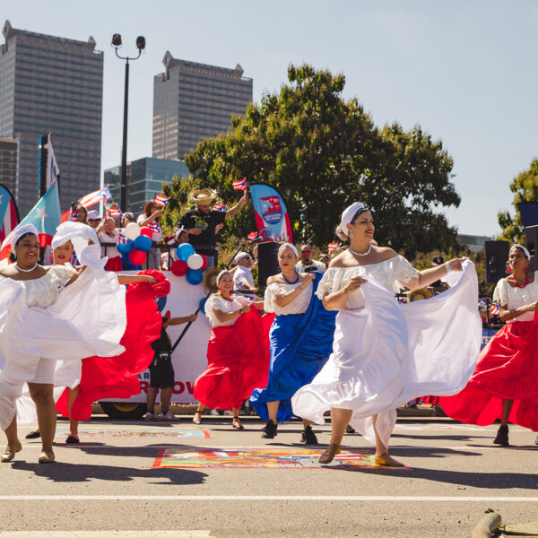 Dancers in flowing red, white and blue dresses perform in the Puerto Rican Day Parade on a car free street as spectators and parade participants gather behind them