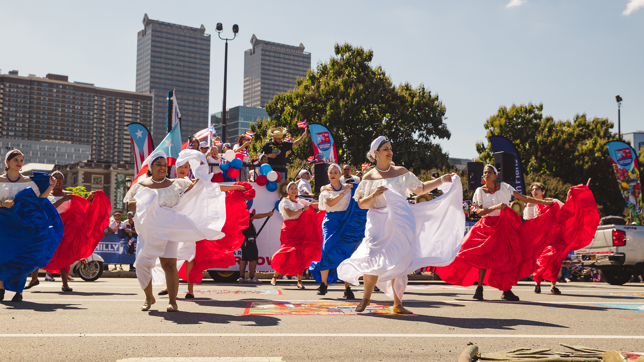 Danseressen in wapperende rode, witte en blauwe jurken treden op tijdens de Puerto Rican Day Parade in een autovrije straat, terwijl toeschouwers en deelnemers aan de parade zich achter hen verzamelen