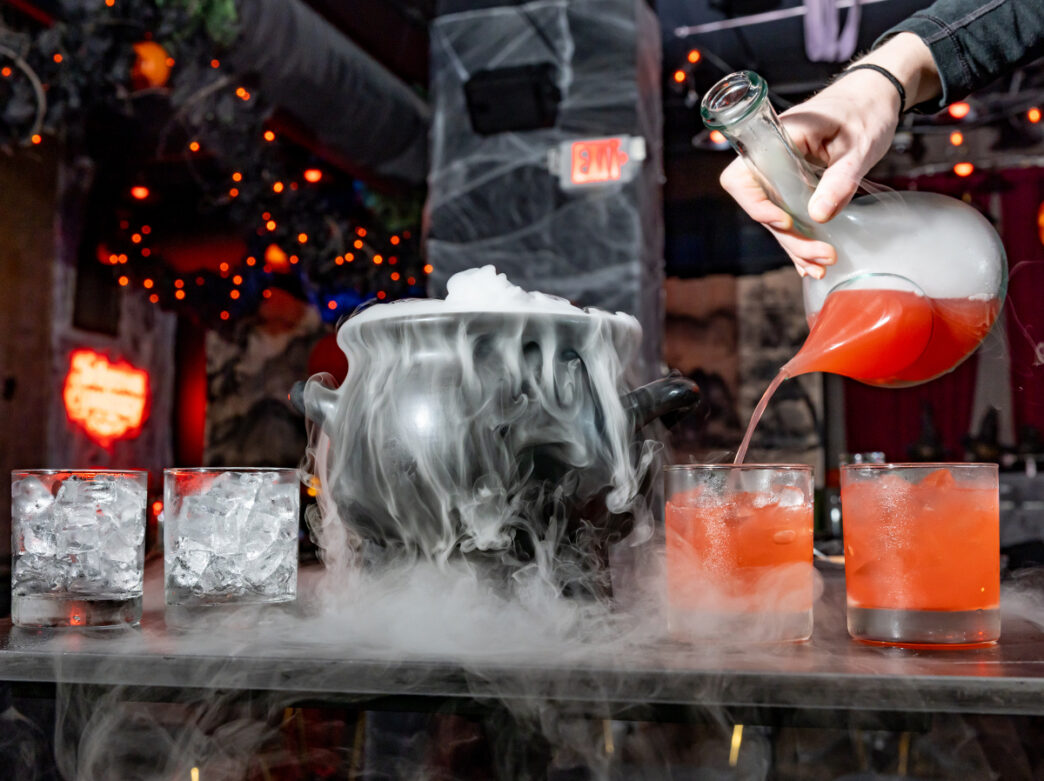 A red cocktail is poured into an ice-filled glasses beside a cauldron overflowing with fog in a festive Halloween setting.