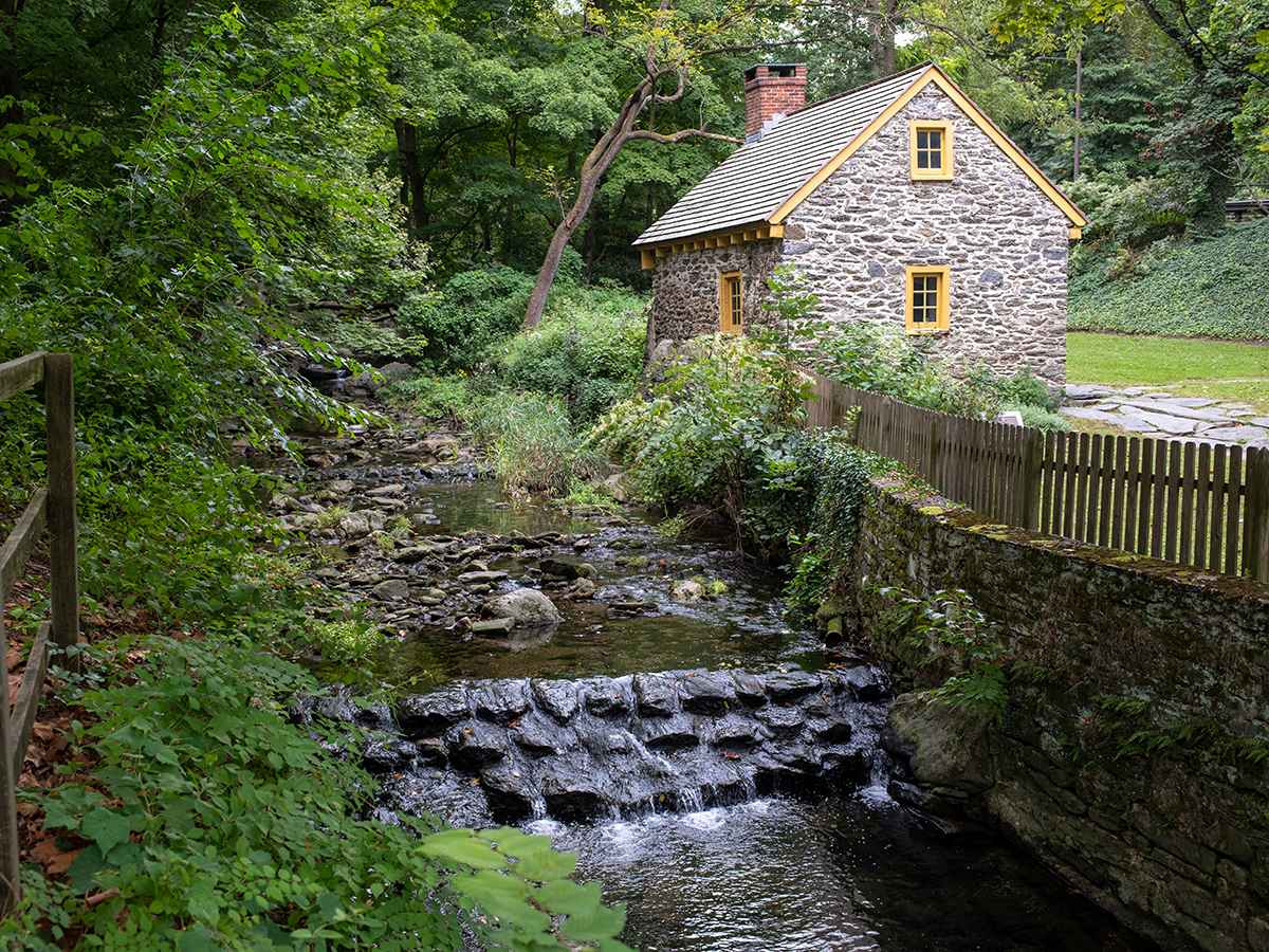 Une maison historique en pierre, aux fenêtres bordées de jaune, se dresse au bord d'un ruisseau rocailleux, au milieu d'une végétation luxuriante, dans le village historique de Rittenhouse.