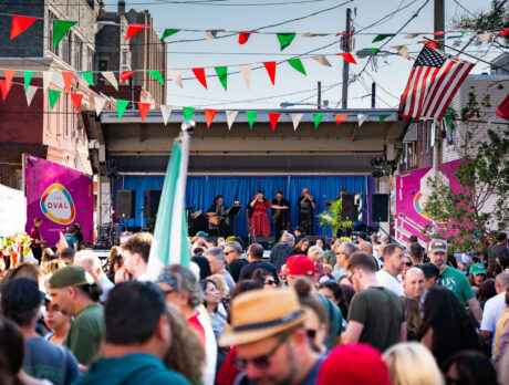 A large crowd fills a neighborhood street during an outdoor Italian festival, with red, white and green pennants overhead and live music onstage.