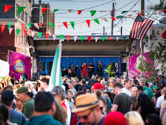 A large crowd fills a neighborhood street during an outdoor Italian festival, with red, white and green pennants overhead and live music onstage.