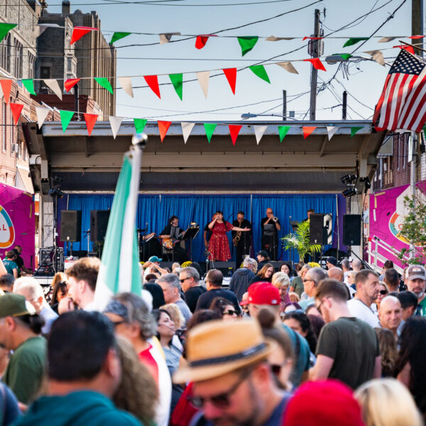 Une foule nombreuse envahit une rue du quartier à l'occasion d'un festival italien en plein air, avec des fanions rouges, blancs et verts suspendus au-dessus des têtes et un concert sur scène.