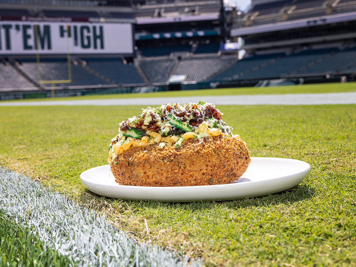 A loaded, crispy-coated baked potato topped with macaroni and cheese, sauce and chopped chicken cutlet sitting on a white plate on the field at Lincoln Financial Field.