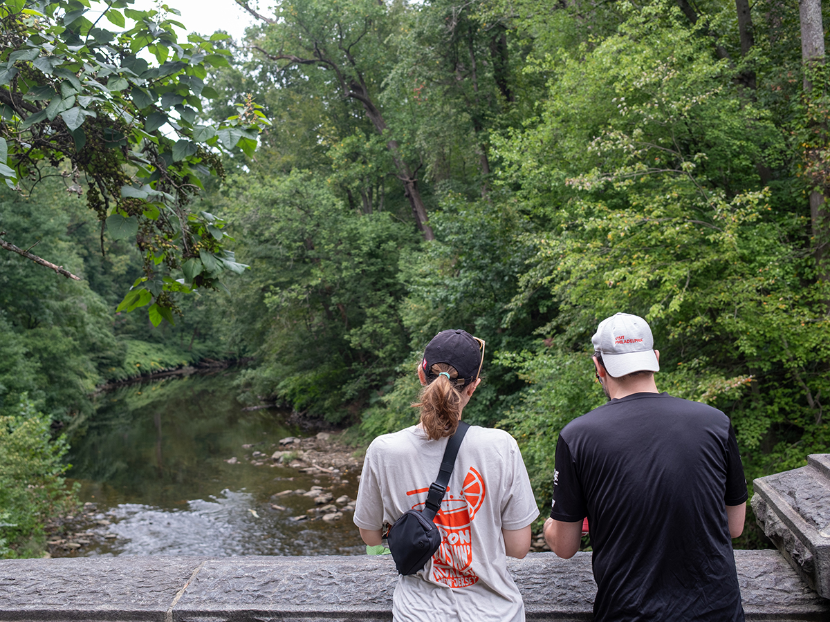 Deux visiteurs se tiennent dos à la caméra sur un pont en pierre surplombant un ruisseau qui serpente à travers une forêt dense et verdoyante, au Wissahickon Valley Park.