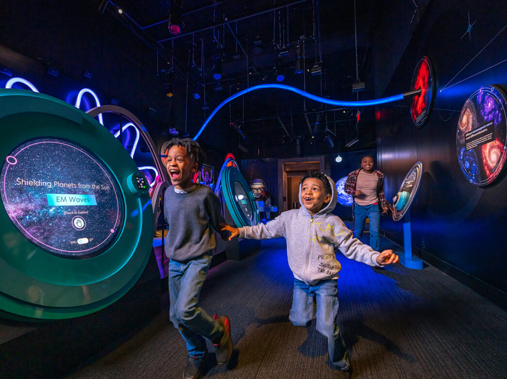 Two excited kids run hand-in-hand through a dark, blue-lit space exhibit filled with colorful interactive displays about planets and wavy LED lights.