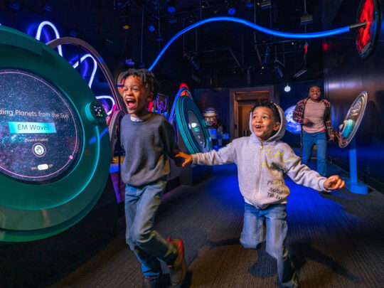 Two excited kids run hand-in-hand through a dark, blue-lit space exhibit filled with colorful interactive displays about planets and wavy LED lights.