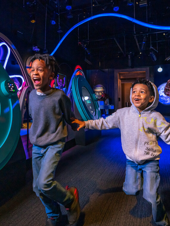 Two excited kids run hand-in-hand through a dark, blue-lit space exhibit filled with colorful interactive displays about planets and wavy LED lights.