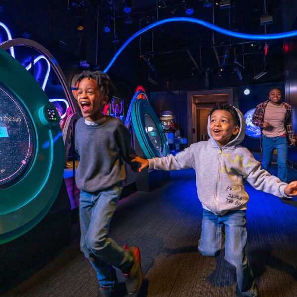 Deux enfants tout excités courent main dans la main à travers une exposition spatiale plongée dans la pénombre et éclairée d'une lumière bleue, où se succèdent des installations interactives colorées sur les planètes et des lumières LED ondulantes.
