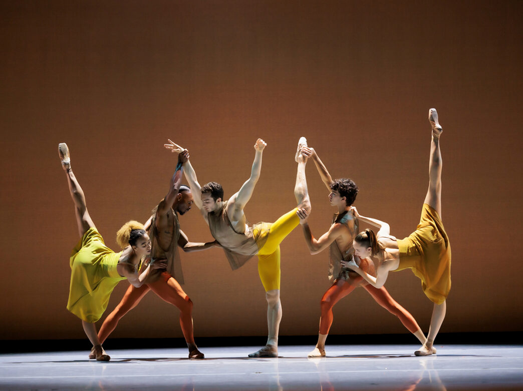 Five ballet dancers in flowing yellow and orange costumes perform a synchronized pose with their legs extended high against a warm brown stage backdrop.