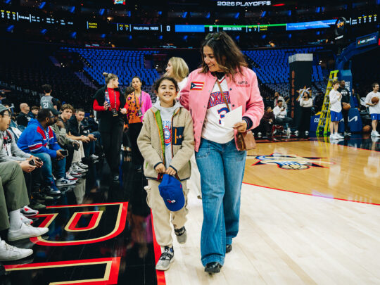 Une femme et un enfant, vêtus de tenues aux couleurs des 76ers de Philadelphie, marchent au bord du terrain à la Xfinity Mobile Arena tandis que les joueurs s'échauffent sur le terrain de basket.