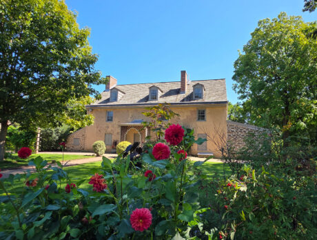 A historic stone house at Bartram’s Garden is surrounded by lush trees, greenery and blooming red flowers on a sunny day.