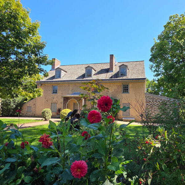 Una casa histórica de piedra en Bartram’s Garden está rodeada de frondosos árboles, vegetación y flores rojas en plena floración en un día soleado.