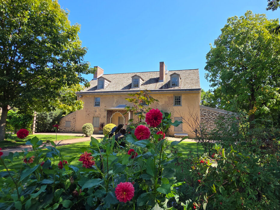 Una casa histórica de piedra en Bartram’s Garden está rodeada de frondosos árboles, vegetación y flores rojas en plena floración en un día soleado.