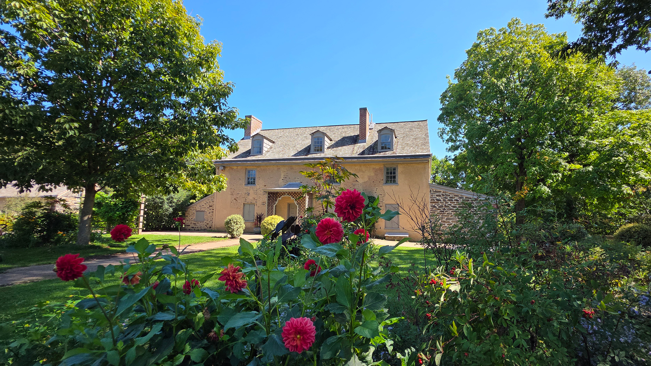 Een historisch stenen huis in Bartram’s Garden wordt op een zonnige dag omringd door weelderige bomen, groen en bloeiende rode bloemen.