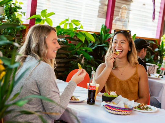 Two women smile and enjoy chips, guacamole, Coca-Cola and other menu items together at a table surrounded by lush greenery inside a colorful restaurant.