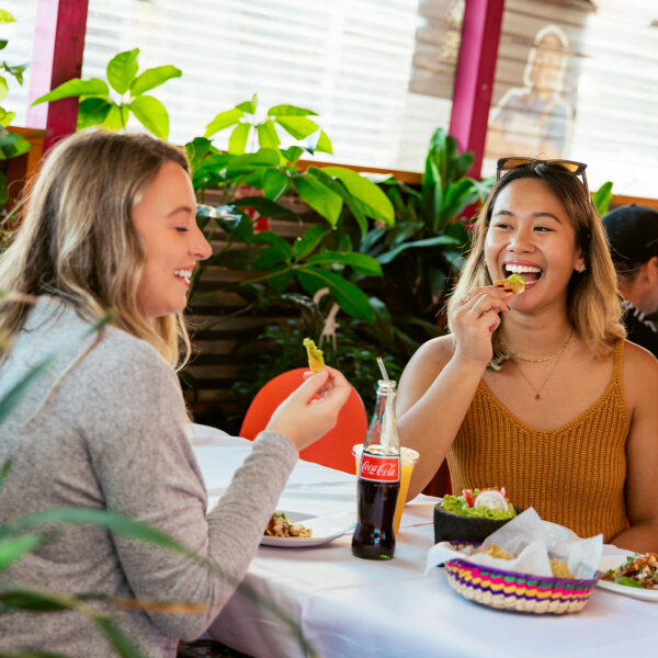 Dos mujeres sonríen y disfrutan juntas de papas fritas, guacamole, Coca-Cola y otros platillos del menú en una mesa rodeada de exuberante vegetación dentro de un restaurante colorido.