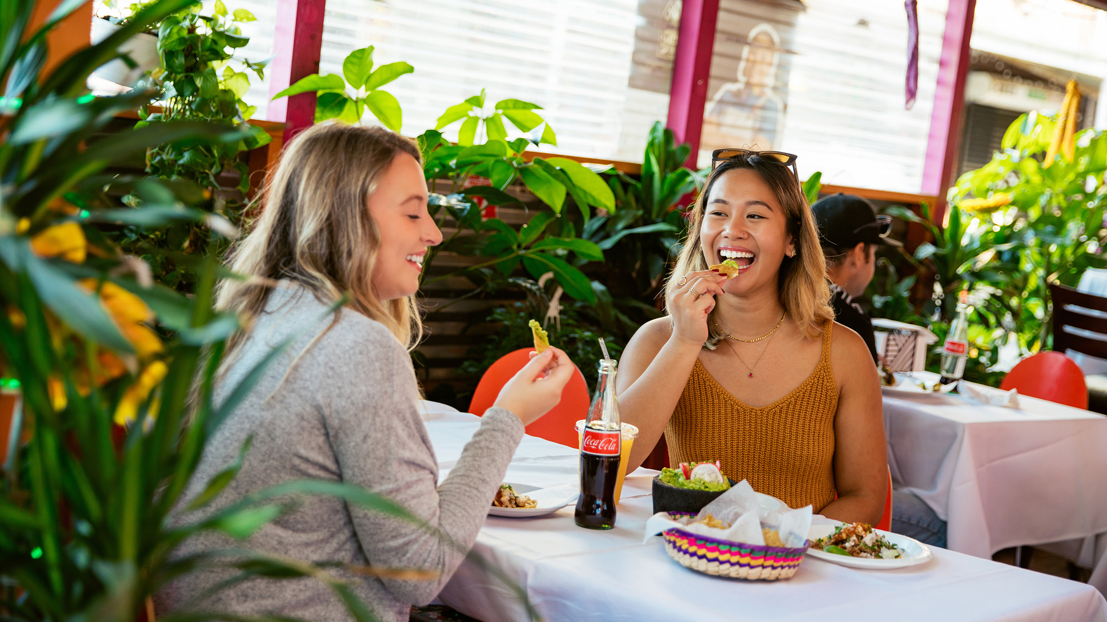 Two women smile and enjoy chips, guacamole, Coca-Cola and other menu items together at a table surrounded by lush greenery inside a colorful restaurant.