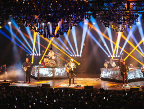 A band performing onstage in front of a packed audience as dramatic blue, white and yellow spotlights crisscross beneath ornate chandeliers.