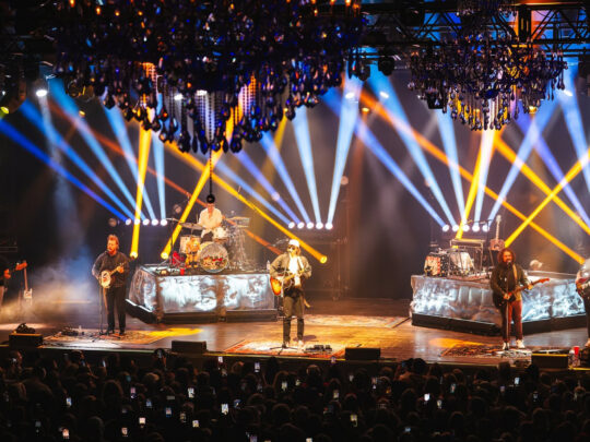 A band performing onstage in front of a packed audience as dramatic blue, white and yellow spotlights crisscross beneath ornate chandeliers.