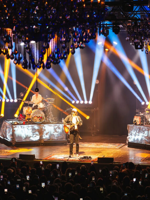 A band performing onstage in front of a packed audience as dramatic blue, white and yellow spotlights crisscross beneath ornate chandeliers.