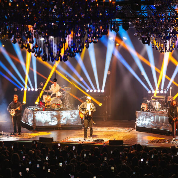 A band performing onstage in front of a packed audience as dramatic blue, white and yellow spotlights crisscross beneath ornate chandeliers.