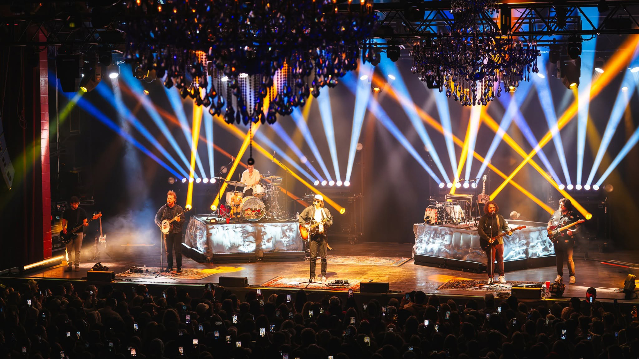 A band performing onstage in front of a packed audience as dramatic blue, white and yellow spotlights crisscross beneath ornate chandeliers.