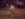 A cowboy in a straw hat and leather cowboy boots rides a bucking horse in an indoor rodeo arena as other cowboys watch from behind.
