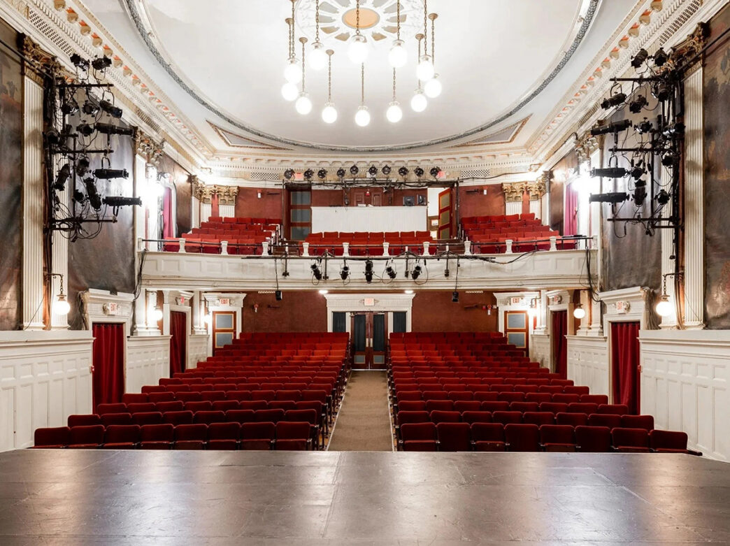 El interior de un teatro histórico con butacas rojas, un balcón superior, detalles arquitectónicos ornamentados y las luces del escenario, visto desde el escenario.