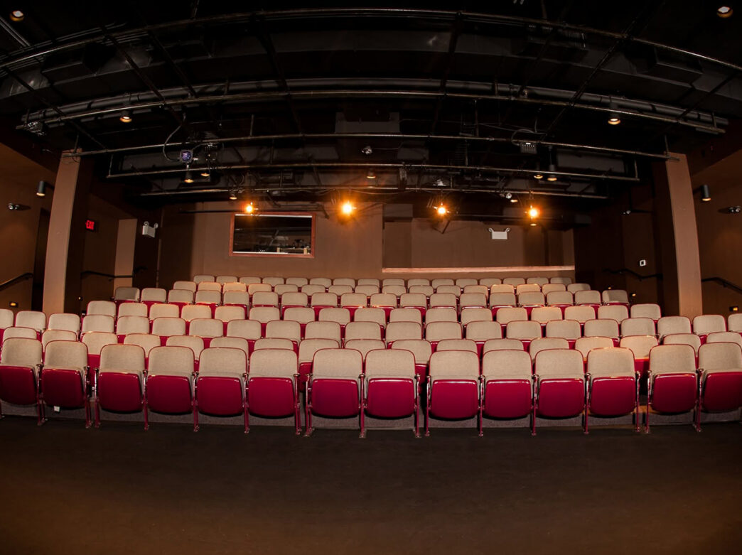Un teatro íntimo, al estilo “caja negra”, con filas de asientos de color beige y rojo orientados hacia el escenario, bajo la iluminación a la vista y las vigas del techo.