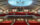 Un auditorio de teatro vacío con hileras de asientos rojos en los niveles de la platea y el balcón, bajo una gran estructura iluminada en el techo.