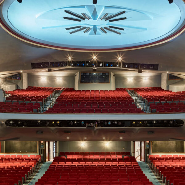 An empty theater auditorium with rows of red seats across the orchestra and balcony levels beneath a large illuminated ceiling feature.
