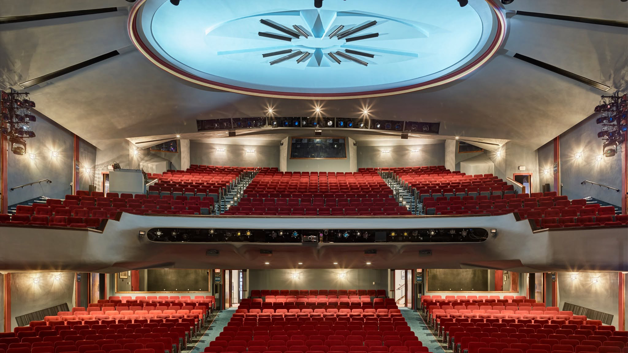 Un auditorio de teatro vacío con hileras de asientos rojos en los niveles de la platea y el balcón, bajo una gran estructura iluminada en el techo.