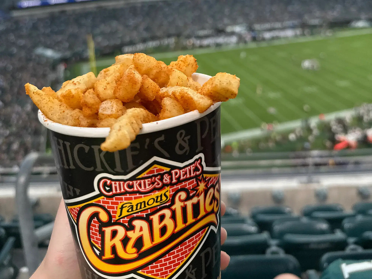 A cup of Chickie's & Pete's seasoned French fries is held in the foreground with a football field and stadium seating in the background.